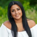 A smiling Hispanic woman with long black hair outdoors in a natural setting, wearing an off-shoulder top.